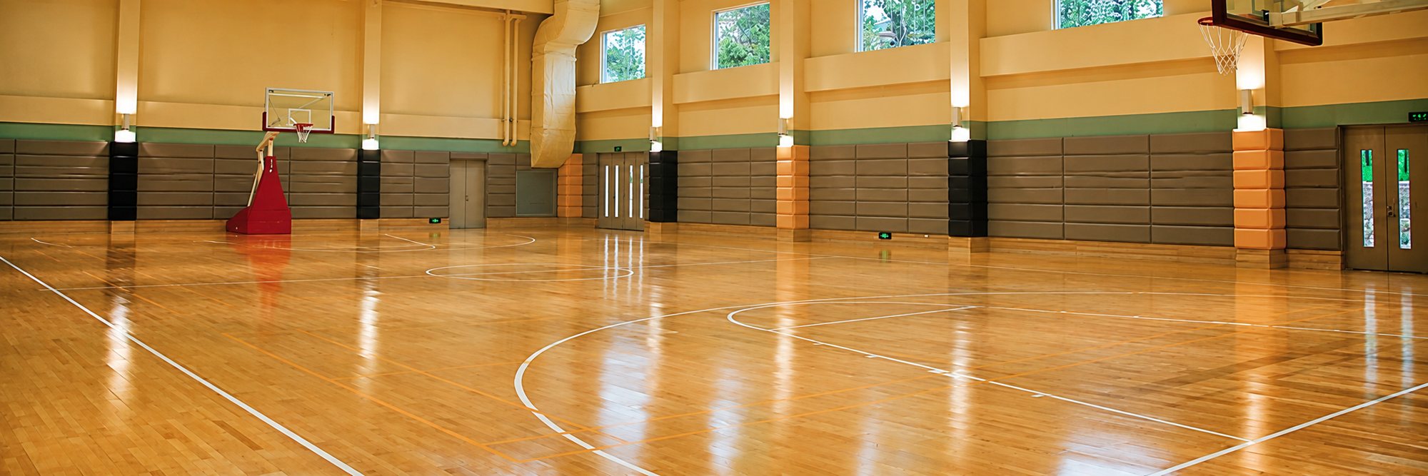 Vaste salle de sport au parquet brillant avec lignes de terrain de basket, paniers et murs jaunes percés de fenêtres.