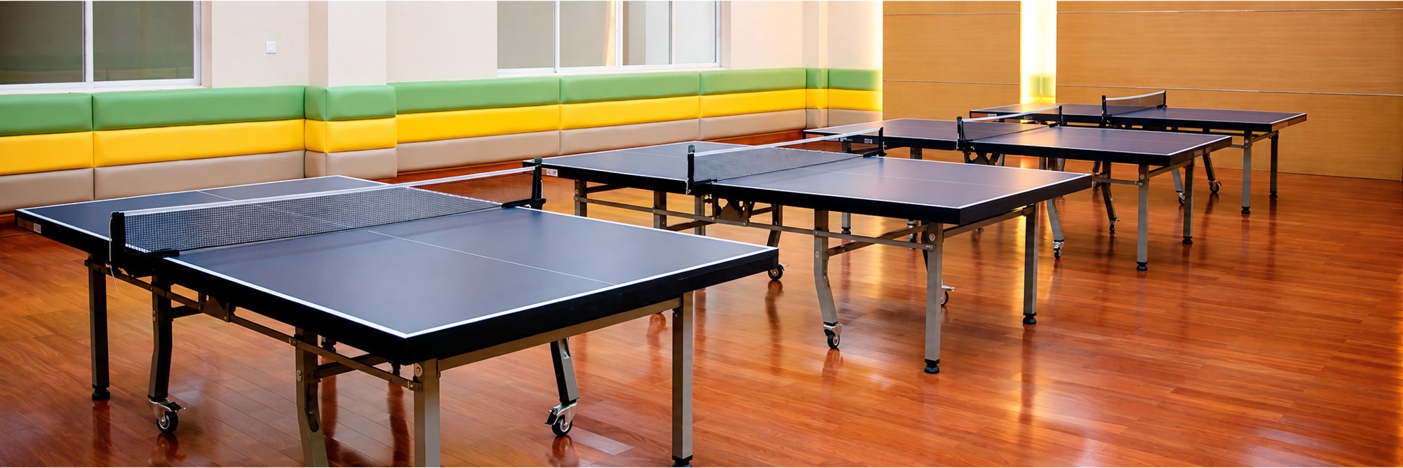 Plusieurs tables de ping-pong bleu foncé alignées dans une grande salle au parquet brillant, avec des banquettes vertes et jaunes le long du mur.