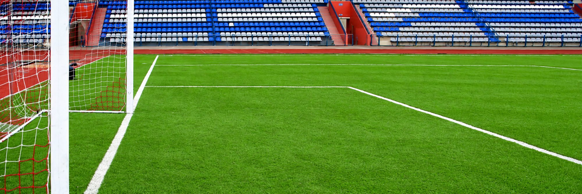 Vue d'un terrain de football en gazon synthétique vert avec un poteau de but, son filet et des tribunes aux sièges bleus et blancs en arrière-plan.